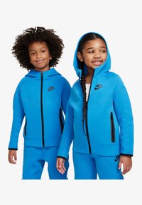 Siniset vetoketjulliset hupulliset takit, jotka ovat pehmeää kangasta ja joissa on mustia yksityiskohtia sekä Nike-logo, yhdistettynä matching sinisiin housuihin.