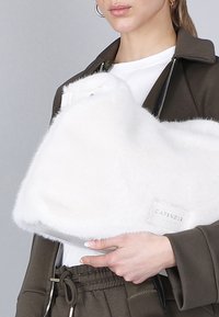 Vrouw in donkere jas en broek die een witte donzige handtas vasthoudt met een zichtbaar "CAFÉNOIR" label op een lichtgrijze achtergrond.