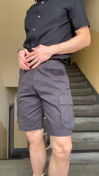 Άνδρας που φοράει μαύρο πουκάμισο με κουμπιά και γκρι σορτς με τσέπες, στέκεται μπροστά από μια σκάλα, τα χέρια του ενώνονται στη μέση.