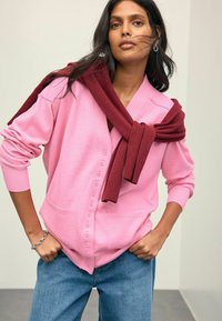 Rosa Strickjacke mit V-Ausschnitt, Knopfverschluss und Seitentaschen, überlagert mit einem burgunderfarbenen Schal, der um die Schultern gebunden ist. Jeans aus Denim.
