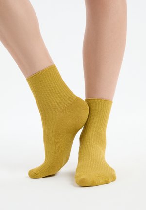 Knöchelhohe, gerippte, senfgelbe Socken, getragen an nackten Füßen, ein Fuß flach aufgesetzt und der andere auf den Zehenspitzen erhoben vor weißem Hintergrund.