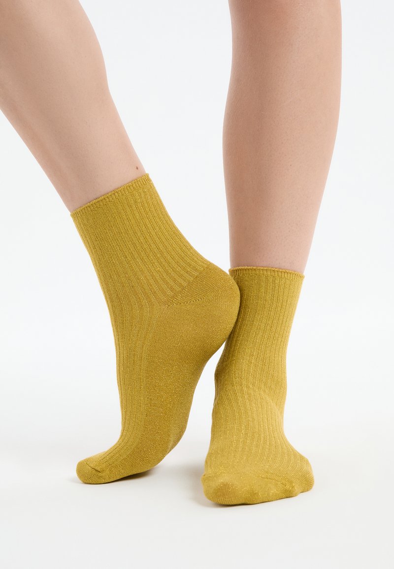 Knöchelhohe, gerippte, senfgelbe Socken, getragen an nackten Füßen, ein Fuß flach aufgesetzt und der andere auf den Zehenspitzen erhoben vor weißem Hintergrund.