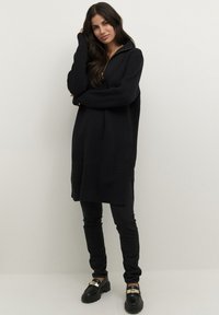 Un pull oversized noir en tricot avec une demi-fermeture éclair, des fentes latérales, porté sur un pantalon noir skinny et des mocassins noirs avec des accents dorés.