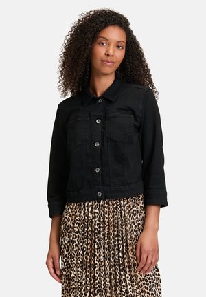 Veste en jean noire cropped avec col, fermeture à boutons et deux poches poitrine. Portée par-dessus une jupe plissée à motif léopard.
