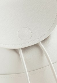 Primo piano di uno zaino bianco in pelle, caratterizzato da una texture liscia, una chiusura a scatto circolare e due sottili cordini. Dettagli di cucitura sottili visibili.
