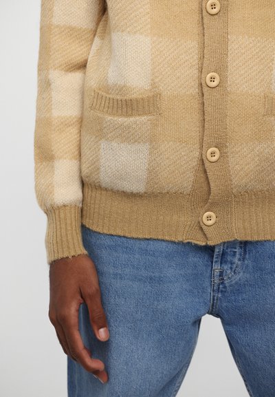 Beige och krämfärgad rutmönstrad cardigan med ribbade mudd och nederkant, med knäppning framtill och två framfickor, tillverkad av mjukt stickat material.