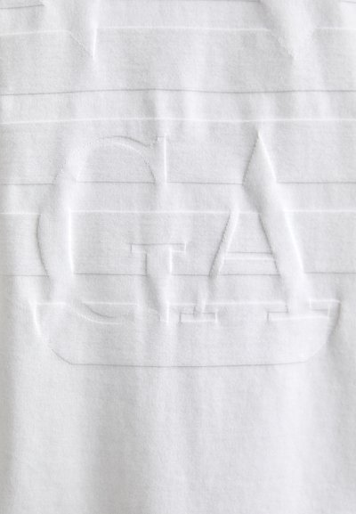 T-shirt en coton blanc avec des rayures horizontales texturées et un logo "GTA" en relief avec un design de bateau sur le devant. Texture douce, style minimaliste.