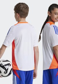 Fehér sportruházatok narancssárga oldalsó panelekkel és kék kiegészítőkkel. Rövid ujjú, kerek nyakkivágású. Két gyerek foci labdával.