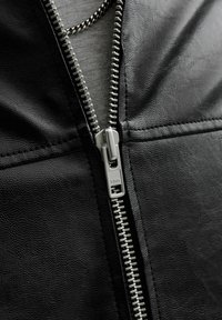 Κοντινό πλάνο ενός ασημένιου μεταλλικού φερμουάρ μισάνοικτου σε ένα μαύρο δερμάτινο μπουφάν με ορατές ραφές και υφή υλικού.