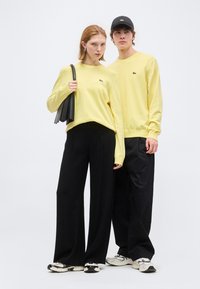 Νεαρός άνδρας και γυναίκα στέκονται κοντά, φορώντας ταιριαστά κίτρινα πουλόβερ, μαύρο παντελόνι με φαρδύ πόδι και λευκά αθλητικά παπούτσια· η γυναίκα κρατά μαύρη τσάντα χειρός.