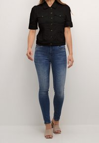 Svart kortärmad cropped skjorta med knäppning och två bröstfickor, matchad med blå skinny jeans och beige öppetåiga skor.