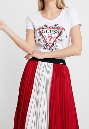 Mujer que lleva una camiseta blanca de Guess con logo floral y una falda plisada de talle alto en paneles rojos y blancos con cinturilla negra.