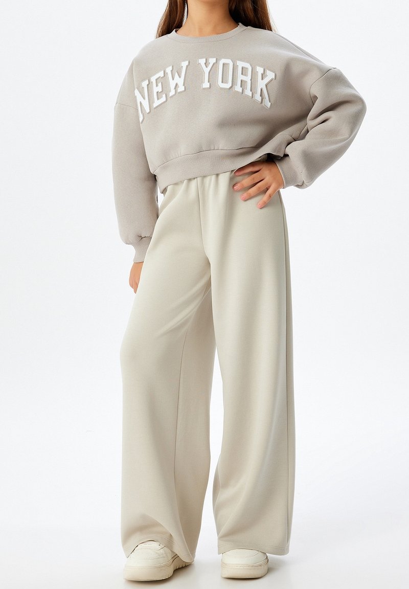 Osoba ubrana w beżowe szerokie spodnie, kremowe trampki oraz taupe'ową bluzę z napisem "NEW YORK" na piersi.