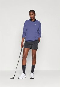 Polo Nike roxa de manga comprida com gola preta, combinada com calções pretos, sapatos brancos e meias azul-marinho, segurando um taco de golfe.