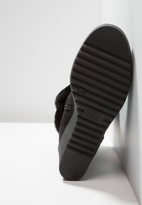 Semelle de botte d'hiver noire avec motif de bande de roulement rainuré, positionnée verticalement contre une surface blanche et un mur.