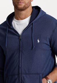 Marinblå zip-hoodie med dragsko, texturerat tyg och en liten vit logotyp på bröstet. Har kängurufickor. Vit t-shirt under.