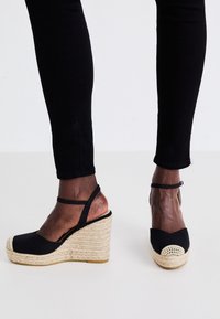 Sandalias de cuña de esparto negras con tacón de yute trenzado, con punta abierta, correa al tobillo y un acento de crochet redondo en la parte frontal.