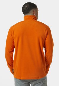 Pomarańczowy polarowy sweter z wysokim kołnierzem i długimi rękawami. Miękka faktura, prosty wzór i metka widoczna na dolnym brzegu. Widok z tyłu.