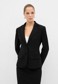 Blazer nero caratterizzato da un design sartoriale, reverse a punta, due tasche frontali e chiusura con un solo bottone, abbinato a una gonna coordinata.