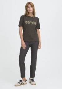 Kobieta stoi ubrana w ciemno-brązowy t-shirt z grafiką, szare dopasowane spodnie i beżowe sneakersy z czarnymi paskami na jednolitym białym tle.
