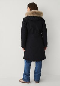Zwarte parka-jas met een met bont gevoerde capuchon, tot de knie, met een taille die is aangesnoerd en van duurzame stof, gecombineerd met blauwe jeans en beige schoeisel.