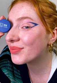 Kremøyenskygge i potte i blått, med et sirkulært design med etikett. Modellen viser en dristig blå eyeliner-design og nøytral leppefarge.