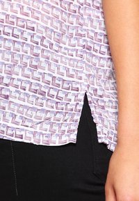 Kortärmad blus i vitt med ett geometriskt mönster i lila, med rundad nederkant och sidofickor. Tillverkad av lätt tyg.