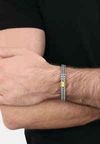 Primer plano de brazos cruzados que llevan una pulsera de cadena plateada con un cierre rectangular dorado en una muñeca peluda de piel clara, vestida de negro.