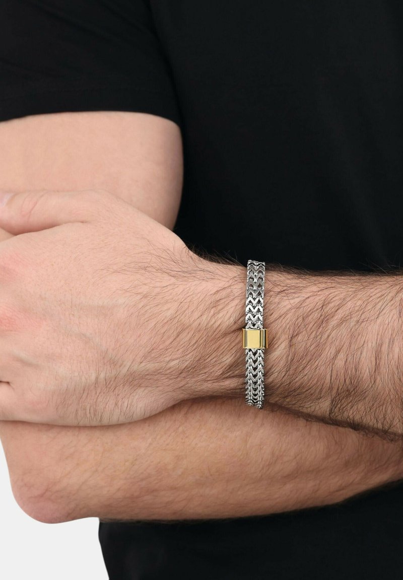 Primer plano de brazos cruzados que llevan una pulsera de cadena plateada con un cierre rectangular dorado en una muñeca peluda de piel clara, vestida de negro.