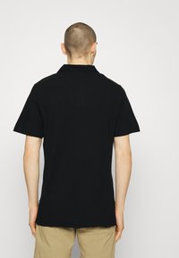 Polo noir à manches courtes, col et coupe droite ; porté avec un short beige clair, affichant un design simple et décontracté.