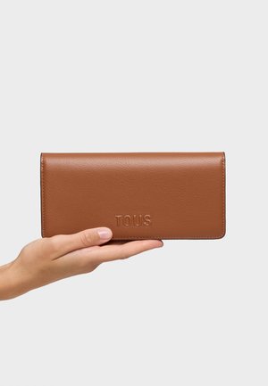 Mano sosteniendo una cartera rectangular de cuero marrón con "TOUS" grabado en el frente sobre un fondo blanco liso.
