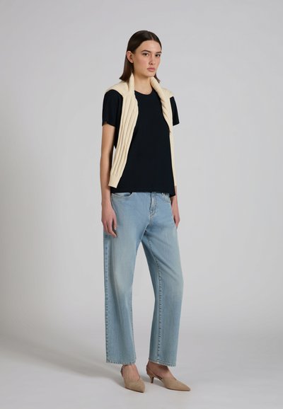 Jonge vrouw staand, gekleed in een zwart T-shirt, lichtblauwe spijkerbroek, beige gebreid vest over de schouders gedrapeerd, en beige pumps met een puntige neus.