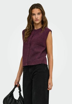 Jeune femme aux longs cheveux ondulés portant un haut violet sans manches boutonné et un pantalon noir, tenant un sac à main noir froncé sur un fond blanc.