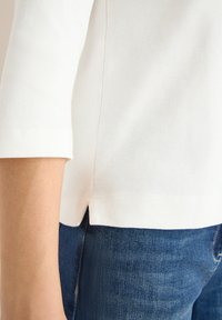 Vit, lättviktig bomullströja med trekvartslånga ärmar, med en rundad nederkant, i kombination med mörkblå denimjeans.