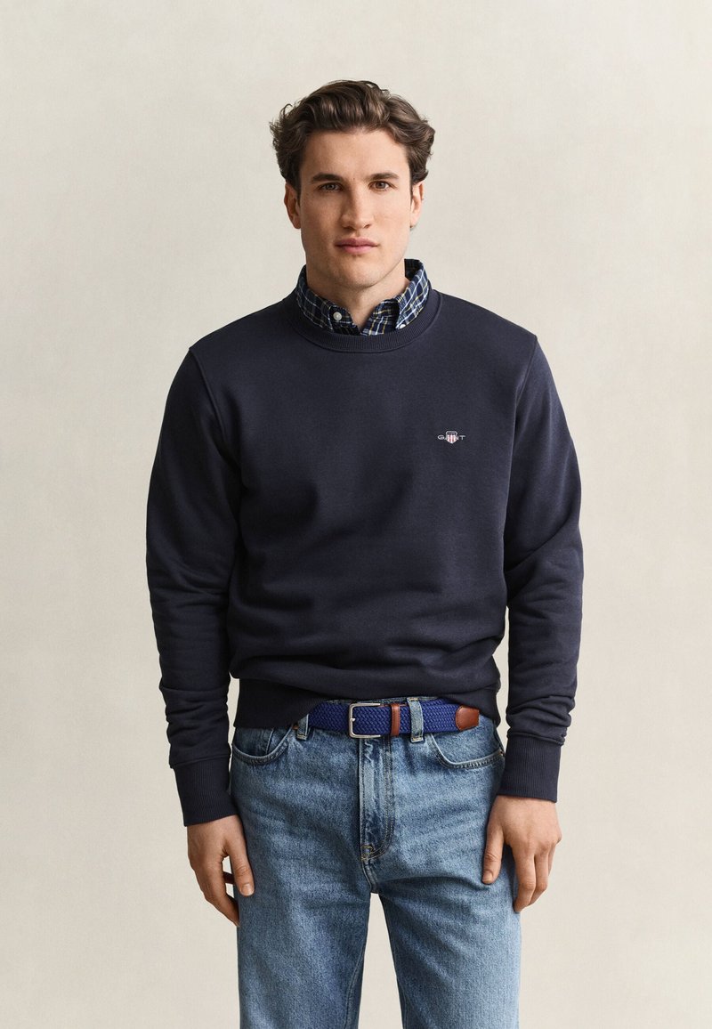 Junger Mann trägt einen marineblauen Pullover über einem karierten Hemd und hellblaue Jeans mit einem gewebten blauen Gürtel und steht vor einem einfarbigen Hintergrund.
