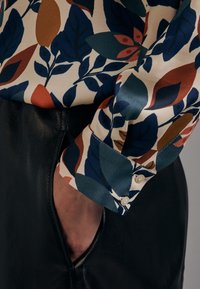 Blouse en soie ornée d'un imprimé feuilles coloré en bleu marine, rouille et sarcelle, avec un poignet boutonné et une jupe en cuir noir. Texture lisse.