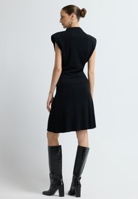 Schwarzes Strickoberteil mit kurzen, strukturierten Ärmeln, kombiniert mit einem passenden, ausgestellten Rock, ergänzt durch knielange, glänzende schwarze Stiefel.