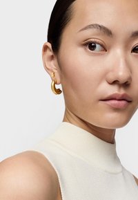 Boucles d'oreilles en or avec un design en forme de vague, associées à une petite puce en or, portées par un mannequin dans un haut texturé de couleur claire.