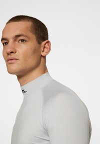 Top grigio chiaro aderente a maniche lunghe con colletto alto e dettagli del logo scuro. Tessuto morbido e design aerodinamico adatto all'uso atletico.