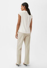 Blouse blanche à manches courtes avec un détail froncé au niveau du col, assortie à un pantalon droit beige clair et des chaussures à talons hauts noires.