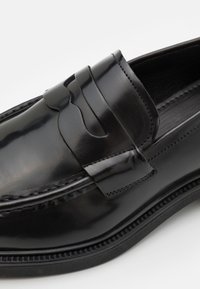 Gros plan d'un mocassin en cuir noir brillant avec une bride décorative et des coutures détaillées sur la tige et la semelle.