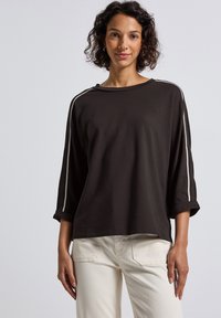 Dunkelbraunes oversized Hemd aus Baumwolle, mit weißen Streifen an den Ärmeln, weitem Ausschnitt und hochgekrempelten Ärmelbündchen, kombiniert mit hellen Cremefarbigen Hosen.