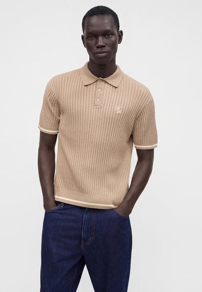 Homme portant un polo en maille beige à manches courtes avec des boutons et un jean bleu marine, debout les mains dans les poches sur un fond uni.