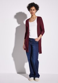 Bordeaux lange cardigan over een witte top, gecombineerd met donkerblauwe flared jeans en lichtgekleurde schoenen. Gladde stof met een getailleerd ontwerp.