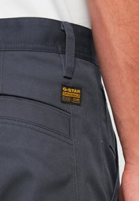 Primo piano di una tasca posteriore di pantaloni grigio scuro con etichetta in denim G-Star Originals Raw gialla e nera e parte di un braccio di una persona.