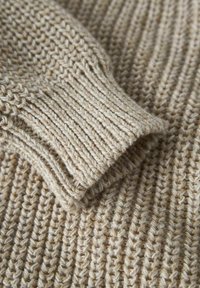 Detail eines beigen Strickpullovers, der die gerippte Textur und das dicke Strickmuster am Bündchen und dem Körperstoff präsentiert.