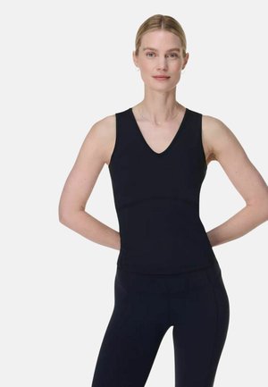 Vrouw met blond haar in een zwarte mouwloze top met V-hals en zwarte broek, staand met handen achter haar rug tegen een effen witte achtergrond.