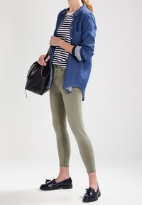 Mujer con camisa de rayas, chaqueta de denim azul, pantalones ajustados oliva, mocasines negros con borlas, sosteniendo un bolso de piel negro.