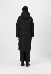 Giacca a vento nera con cappuccio, caratterizzata da un design lungo, un motivo a zig-zag e maniche ampie. Abbinata a pantaloni neri a gamba larga e stivali.