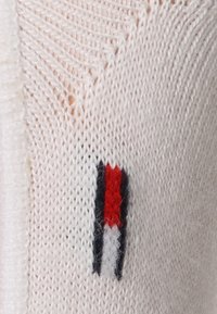 Hvit strikket genser med et rødt, marineblått og hvitt stripete logo-patch på venstre side. Myk tekstur med åpen vev.